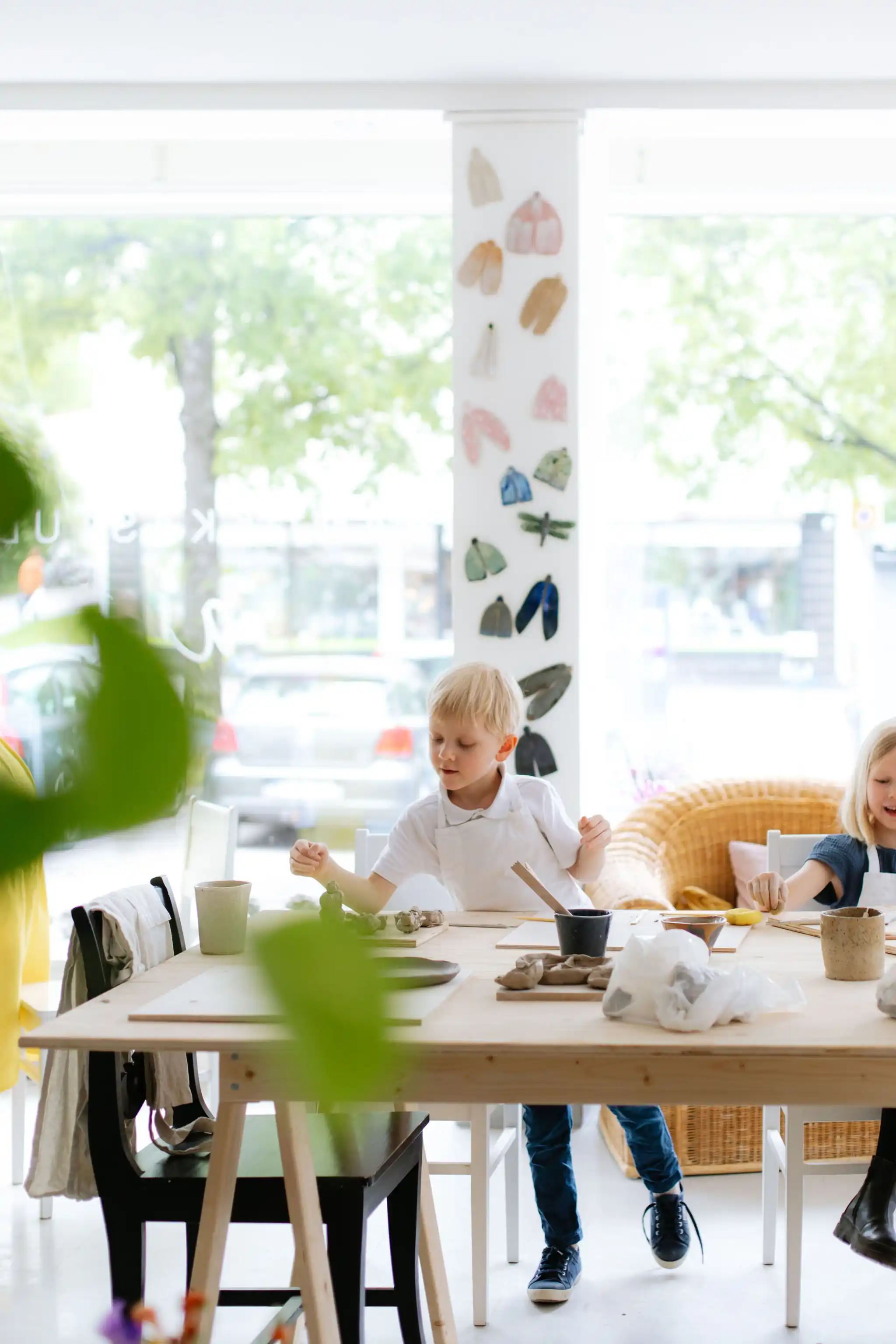 Keskittynyt lapsi muotoilee savea viihtyisässä ja valoisassa keramiikkastudiossa. Pöydällä on työkaluja ja materiaaleja. Ikkunanpielen pylväässä on uniikkeja keramiikkakoristeita, ja suuri ikkuna avautuu vehreälle kadulle.